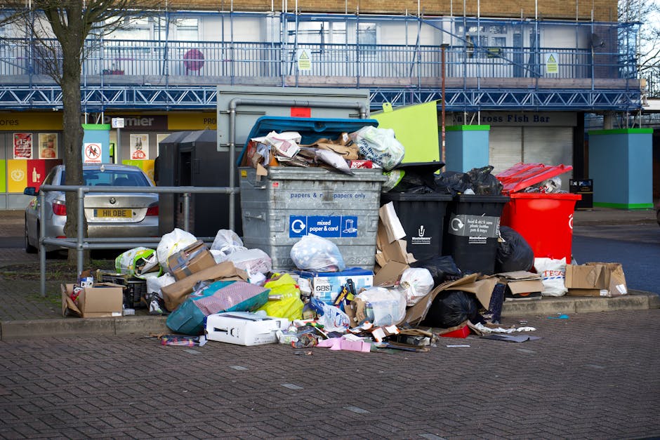 A photograph of a large pile of mixed waste materials piled on an outdoor driveway or paved area. The waste includes various types of discarded items such as cardboard boxes, plastic containers, paper